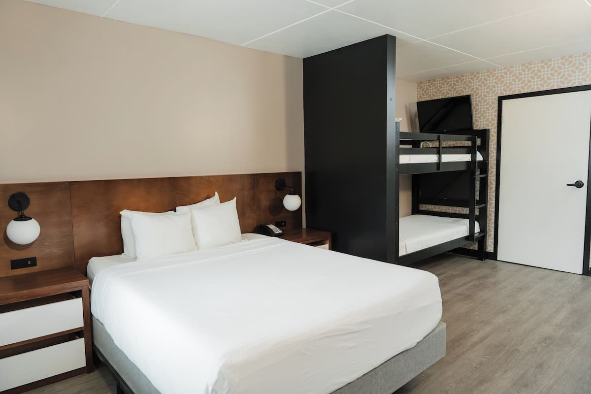 A hotel room with a large bed, a wooden headboard, a nightstand with a lamp, and a built-in bunk bed.