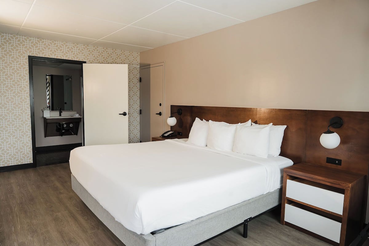 A hotel room with a large bed, a wooden headboard, a nightstand with a lamp, and a bathroom with a mirror and sink.