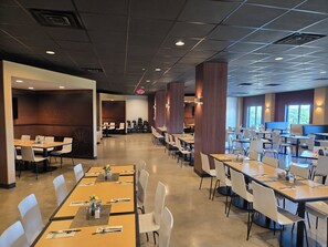 A spacious cafeteria with multiple dining tables and chairs, a central walkway, and large windows providing natural light.