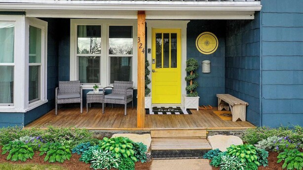A house with a bright yellow door, a porch with wicker furniture, and a garden with various plants.