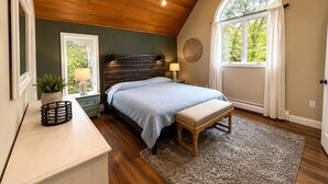 A bedroom with a sloped ceiling, a bed with a headboard, a bench, a nightstand with a lamp, a window with curtains, and a view of trees outside.