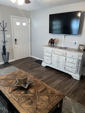 A living room with a wooden coffee table, a white cabinet, a flat-screen TV mounted on the wall, and a door with a coat rack.