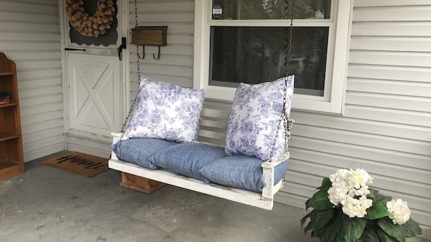 Une véranda avec une balançoire blanche, un banc en bois, une couronne décorative et une plante en pot.
