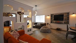 Ett modernt vardagsrum med en soffa, ett soffbord och en tv. Det finns hyllor med dekorativa föremål och en matplats med ett bord och stolar.