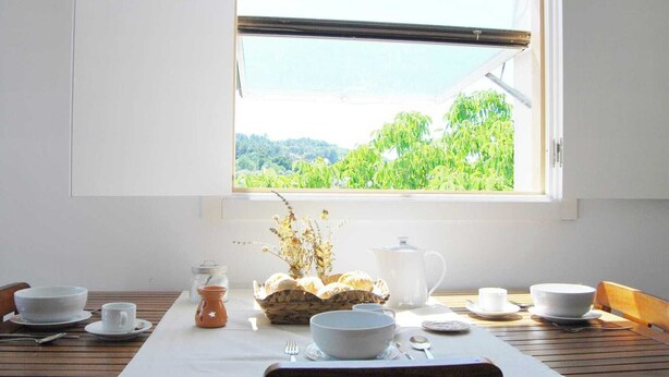 Une table à manger recouverte d’une nappe blanche, agrémentée de bols, de tasses et d’un panier de pain. Une fenêtre donnant sur un paysage verdoyant se trouve au-dessus de la table.