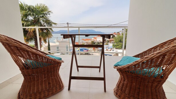 Ein Balkon mit Korbstühlen und einem kleinen Tisch, mit Blick auf eine Küstenlandschaft mit Gebäuden und Palmen.