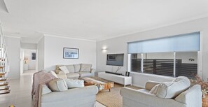 Een moderne woonkamer met een hoekbank, een flatscreen-tv en een groot raam met zonwering.