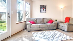 Una sala de estar con un sofá gris, dos cojines rojos, un tapete blanco estampado y un cuadro en la pared.