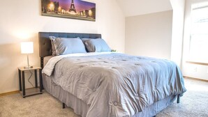 Un dormitorio con una cama grande, una mesita de noche con una lámpara, un cuadro de la Torre Eiffel y una ventana con luz natural.