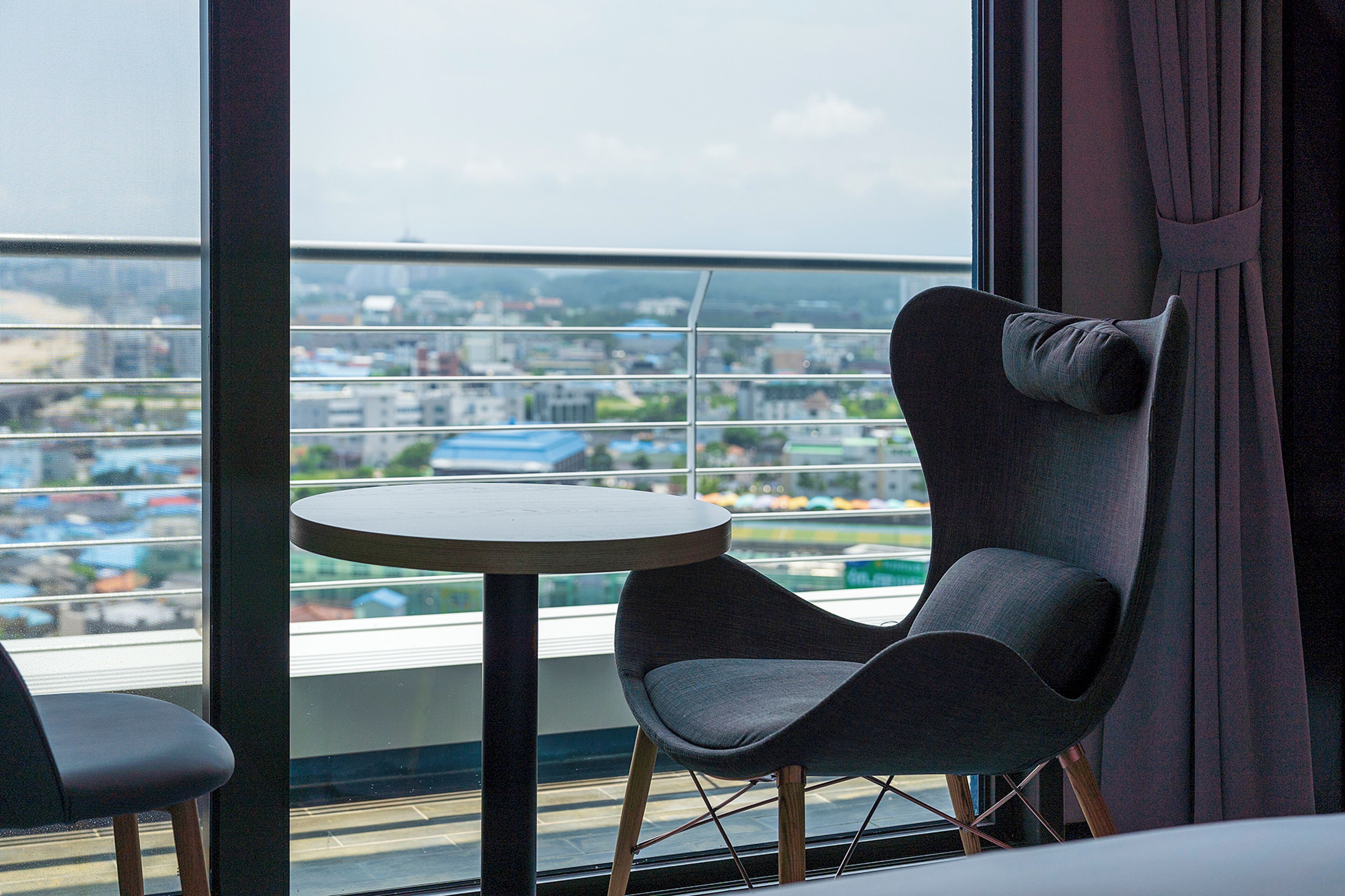Ein modernes Zimmer mit einem runden Tisch und zwei Stühlen, einem großen Fenster mit Blick auf eine Stadtlandschaft und einem Vorhang auf der rechten Seite.