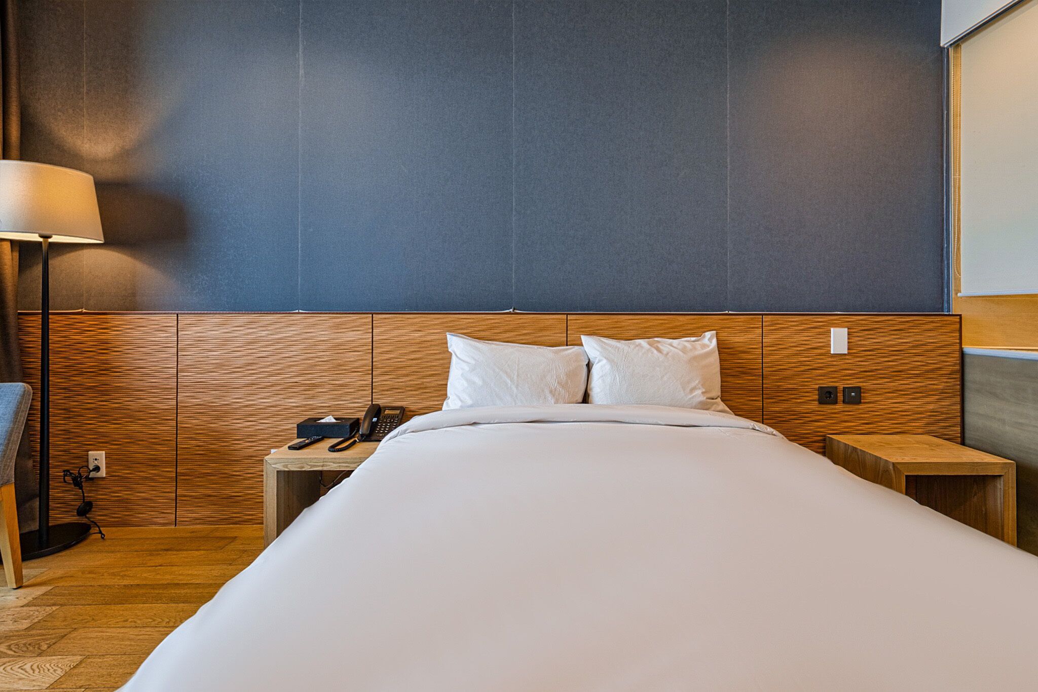 A hotel room with a wooden headboard, a bed with white linens, a bedside table with a phone and a lamp, and a nightstand with a lamp.