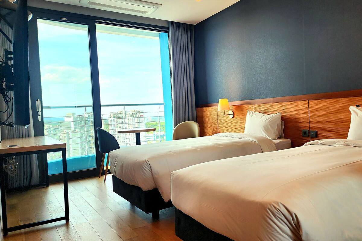 Een hotelkamer met twee bedden, een bureau en een stoel. Er is een groot raam met uitzicht op gebouwen.