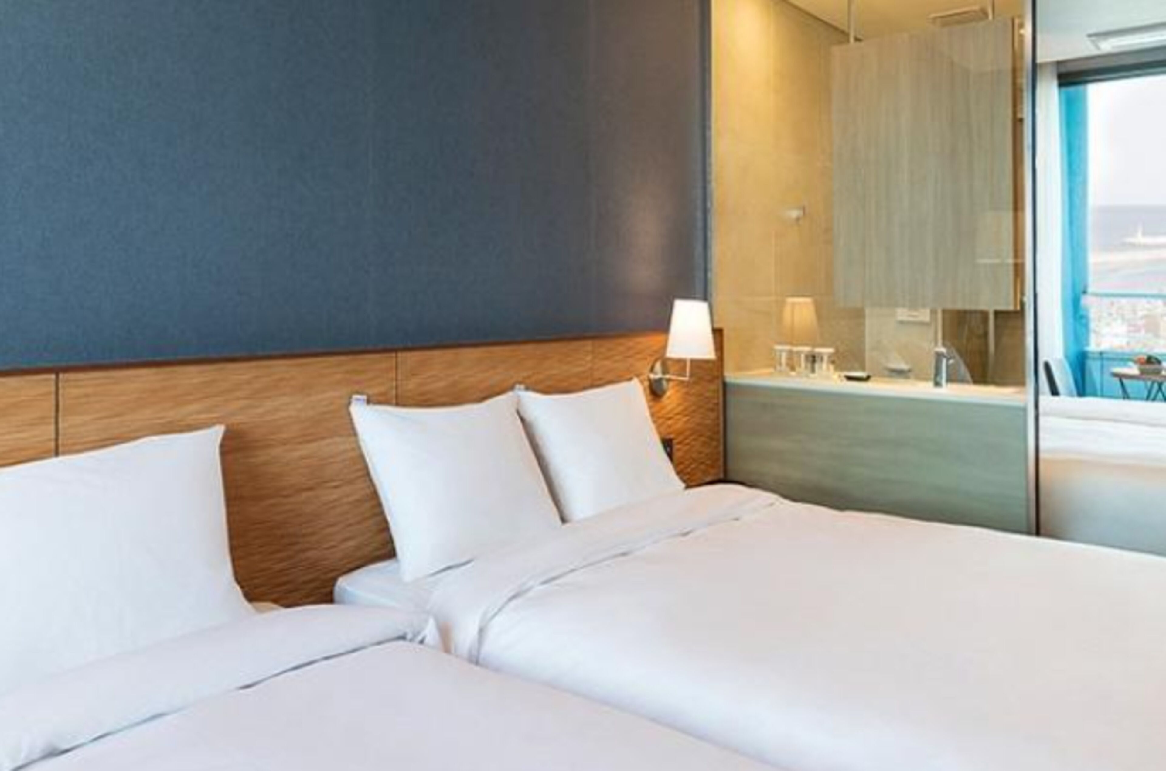 A hotel room with a large bed, a wooden headboard, a nightstand with a lamp, and a view of the city through a large window.