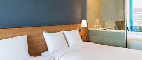 A hotel room with a large bed, a wooden headboard, a nightstand with a lamp, and a view of the city through a large window.