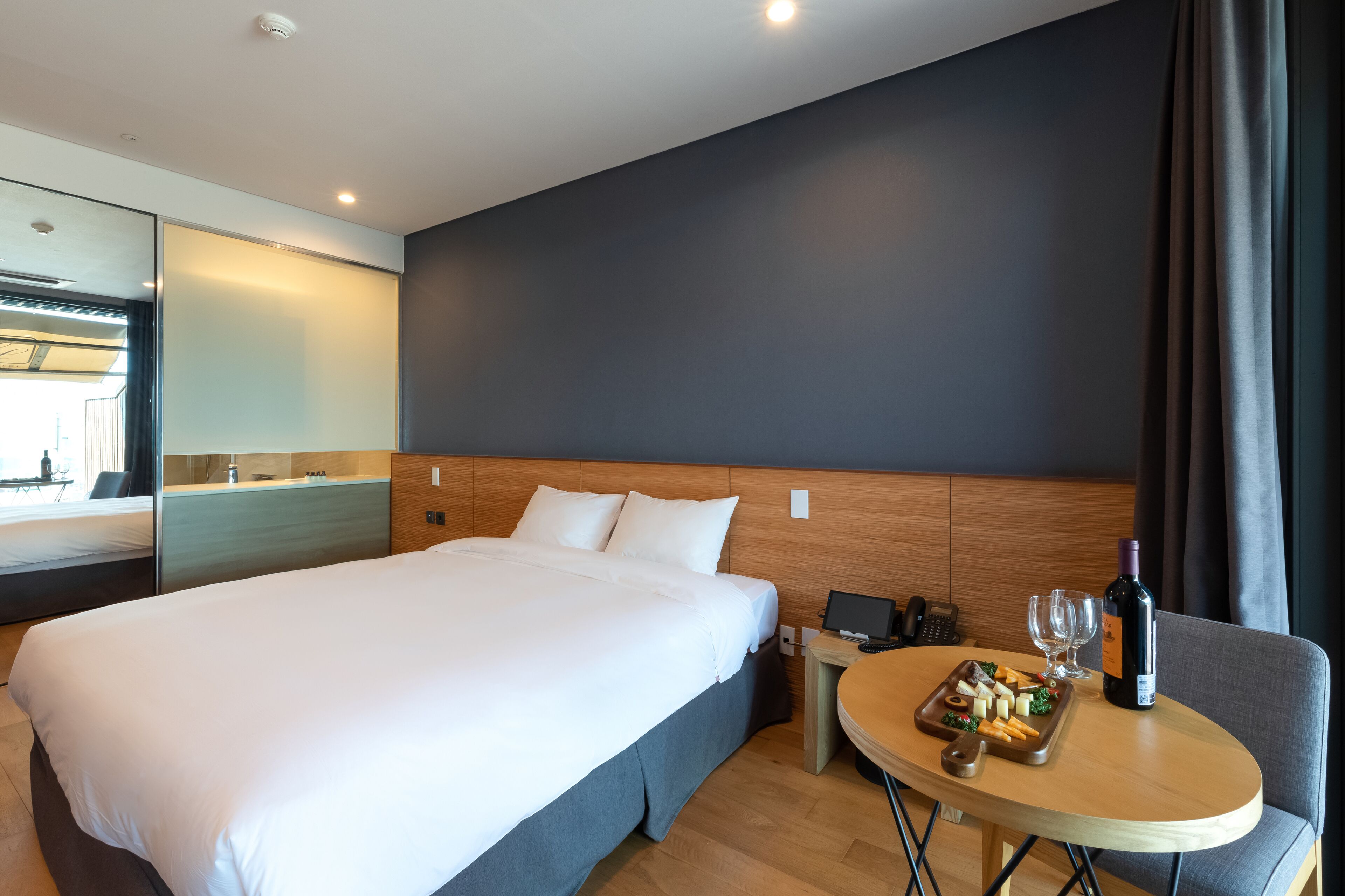 Una habitación de hotel moderna con una cama grande, una mesita de noche con una botella de vino, vasos y una bandeja de aperitivos.