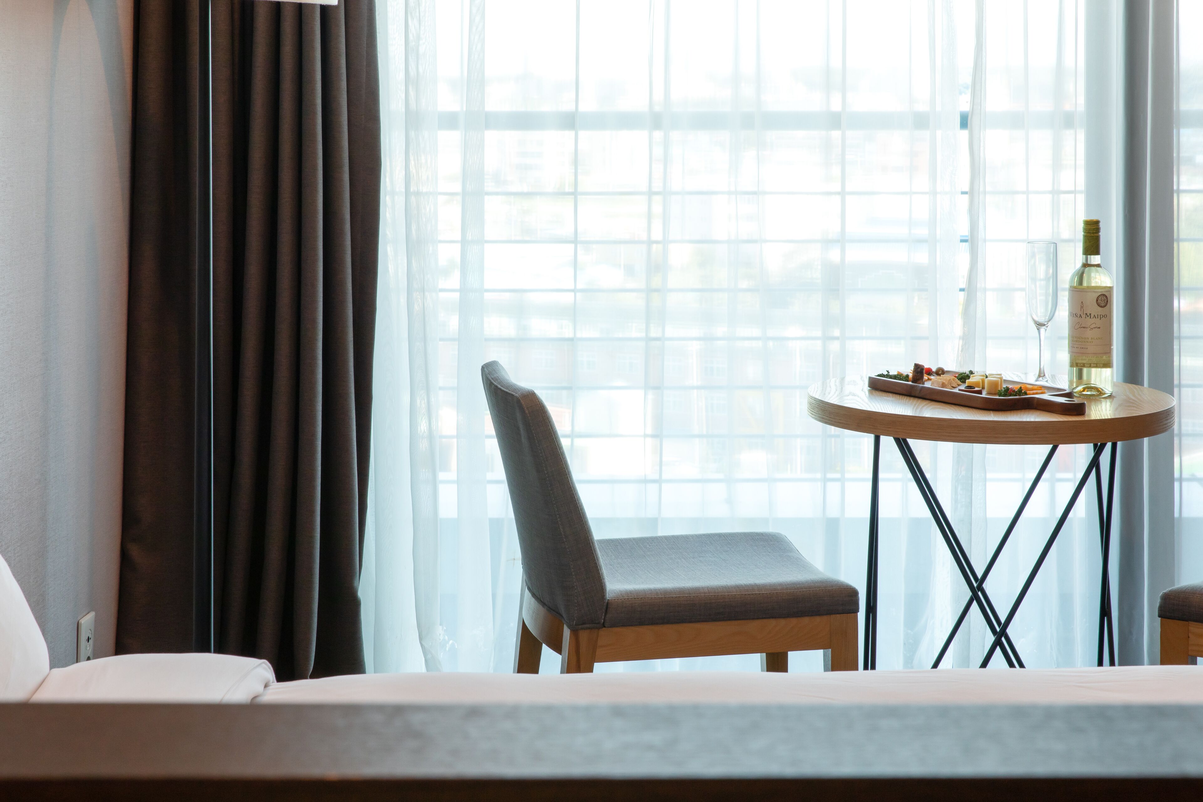 A hotel room with a bed, a chair, a table with a bottle of wine and glasses, and a window with curtains.