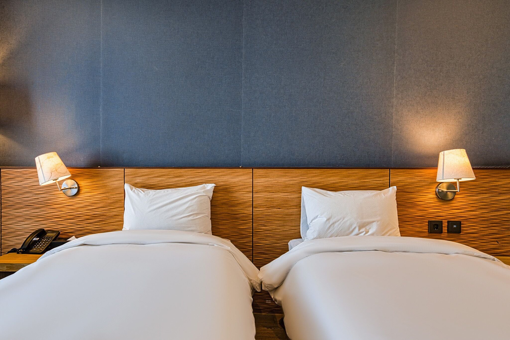 A hotel room with two beds, wooden headboard, bedside lamps, and a phone on the nightstand.