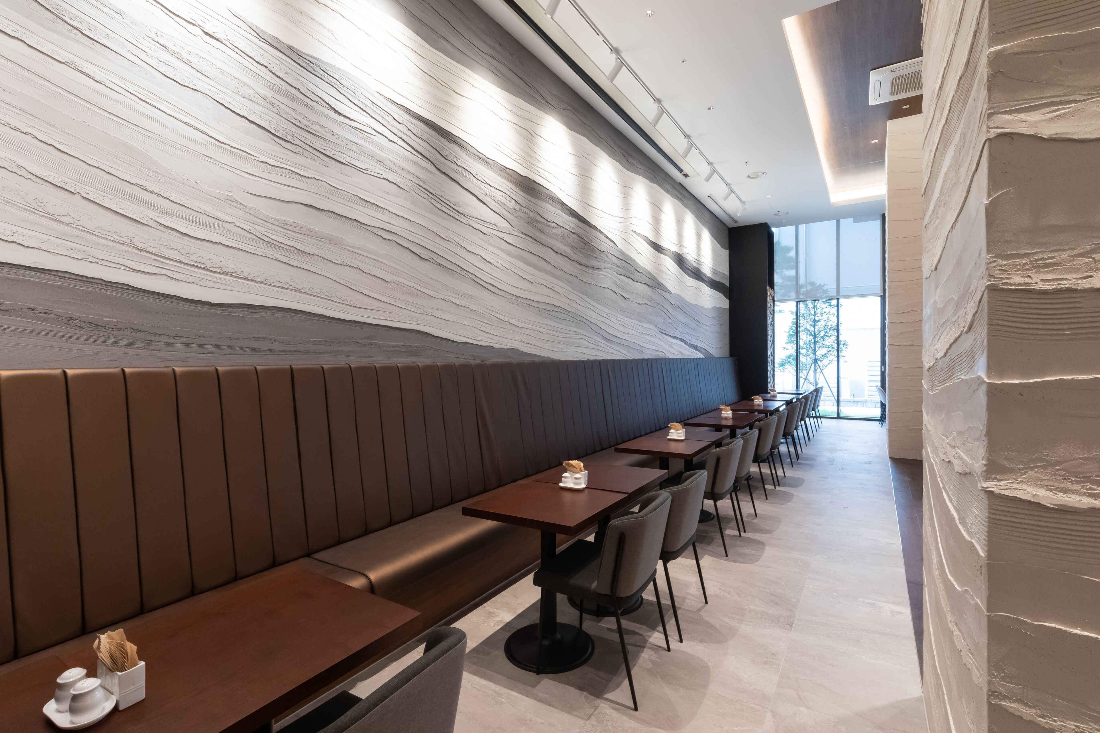 A modern restaurant interior with long wooden tables, cushioned benches, and a unique textured wall.