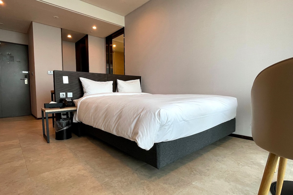 Een moderne hotelkamer met een groot bed, een nachtkastje met telefoon en prullenbak, en een stoel.