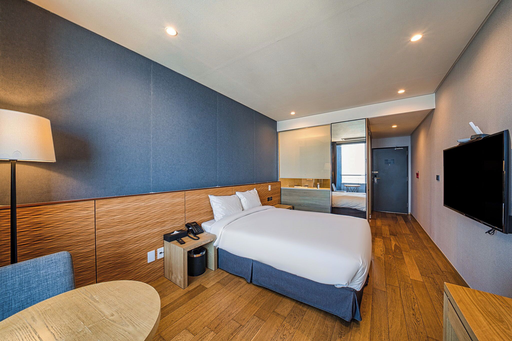 Quarto de hotel moderno com uma cama grande, mesa de cabeceira, abajur e televisão.