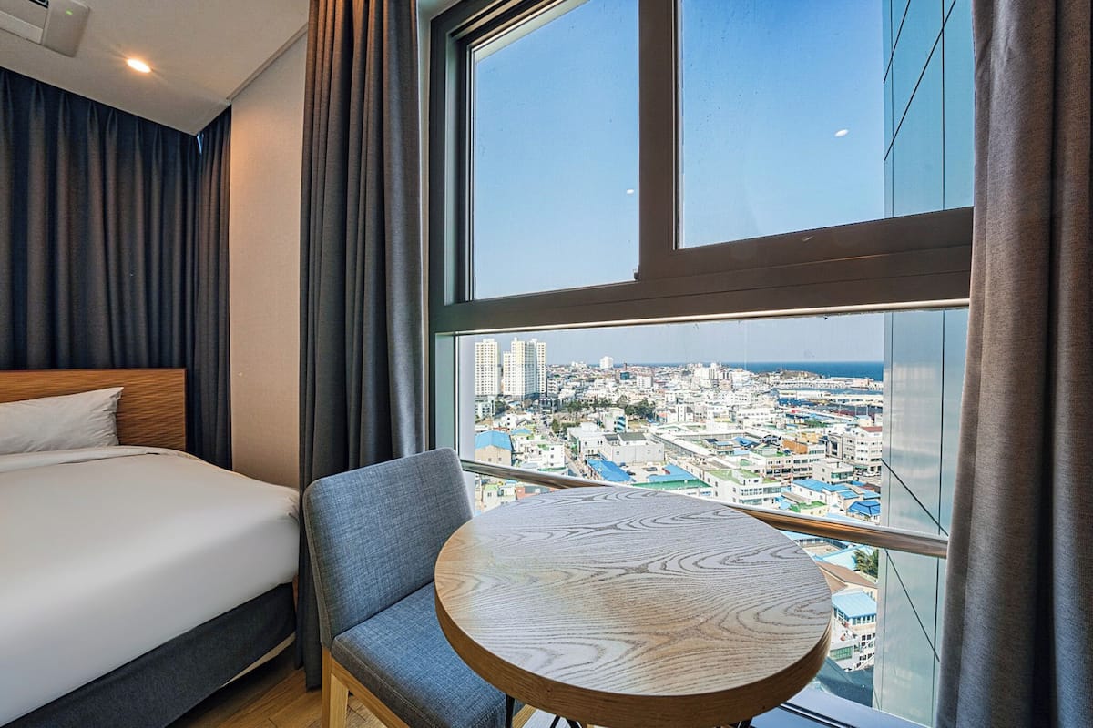 Een hotelkamer met een bed, een stoel, een ronde tafel en een groot raam met uitzicht op de stad.