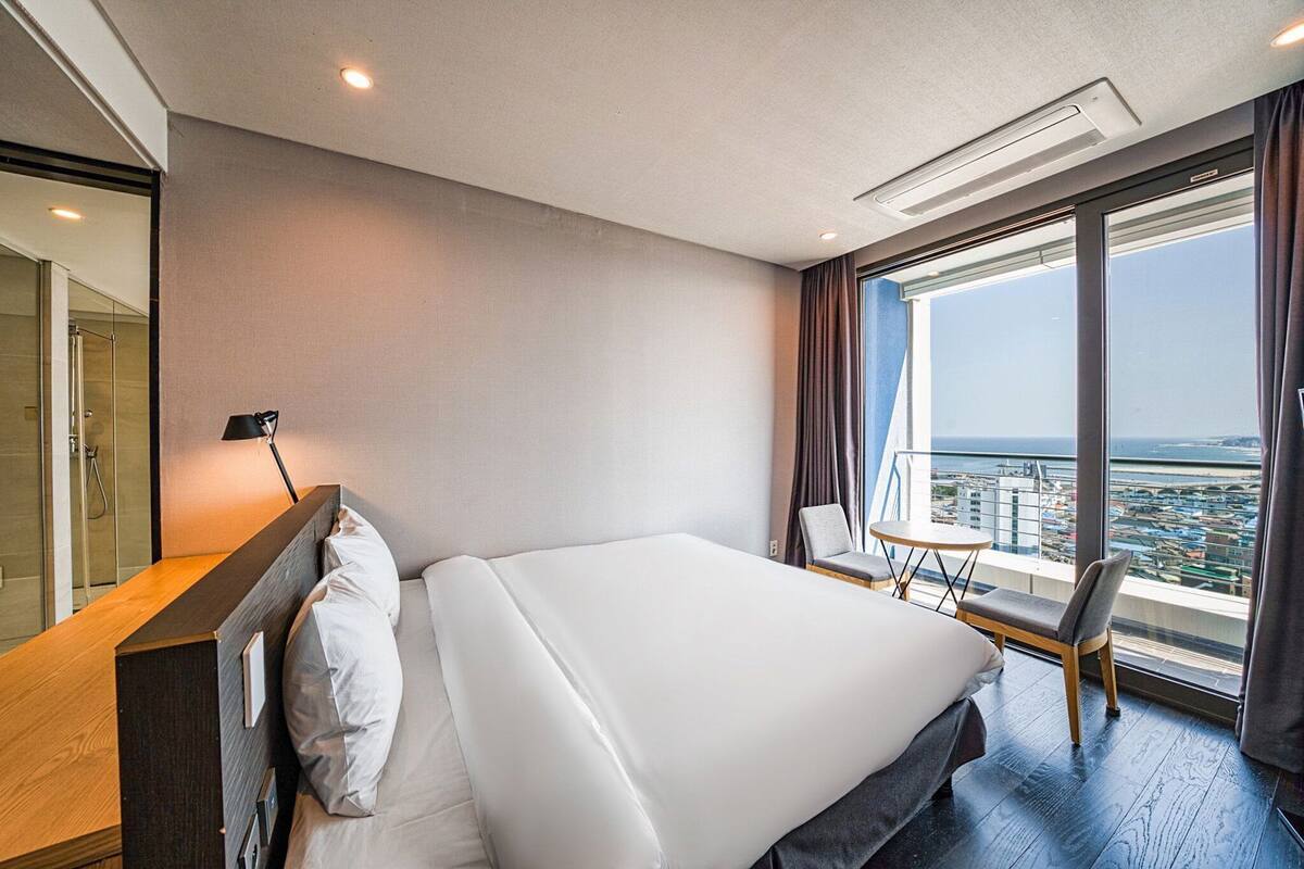 Een moderne hotelkamer met een groot bed, een bureau, een stoel en stadszicht door grote ramen.