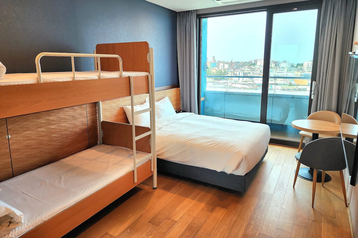 Een hotelkamer met een stapelbed, een eenpersoonsbed, een tafeltje en uitzicht op de stad door een groot raam.