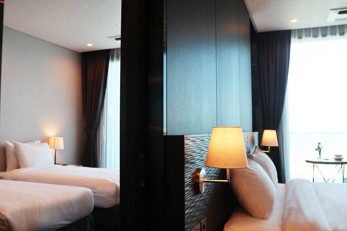 Hotelkamer met twee bedden, een wandlamp, een tafeltje met een kopje en uitzicht op zee door een raam.