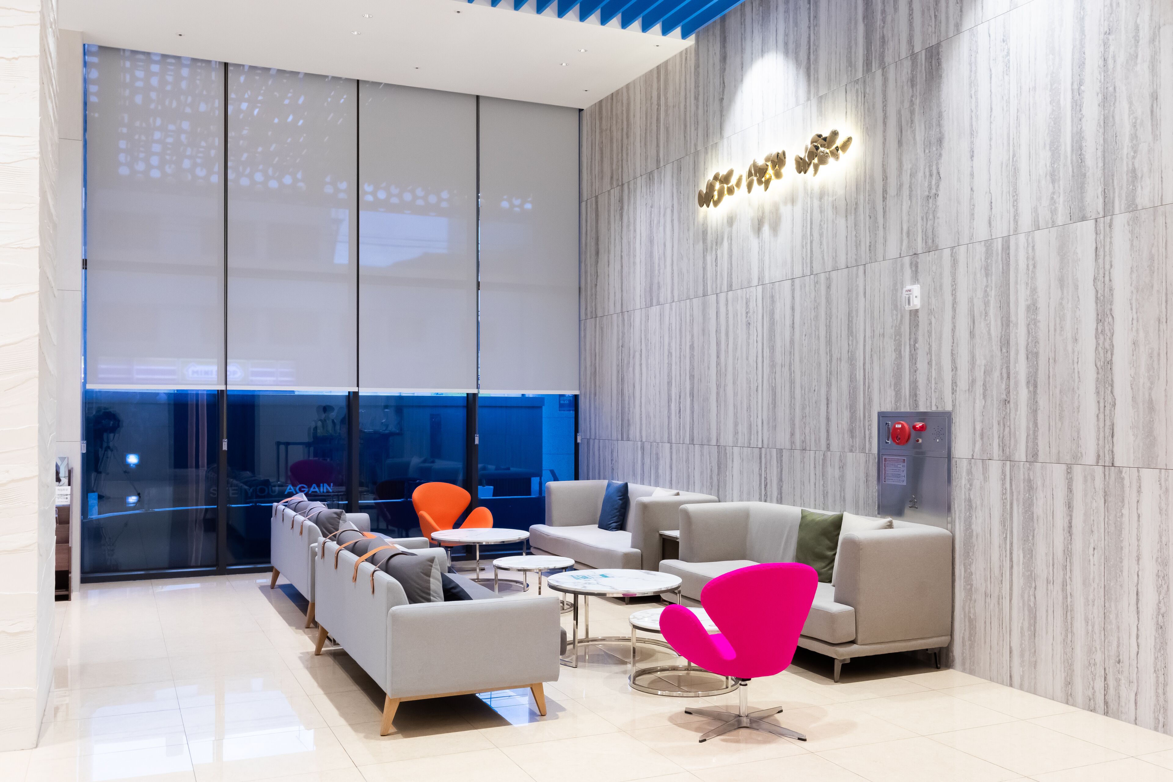 A modern lounge area with sofas, a bright orange chair, and a glass wall with a city view.
