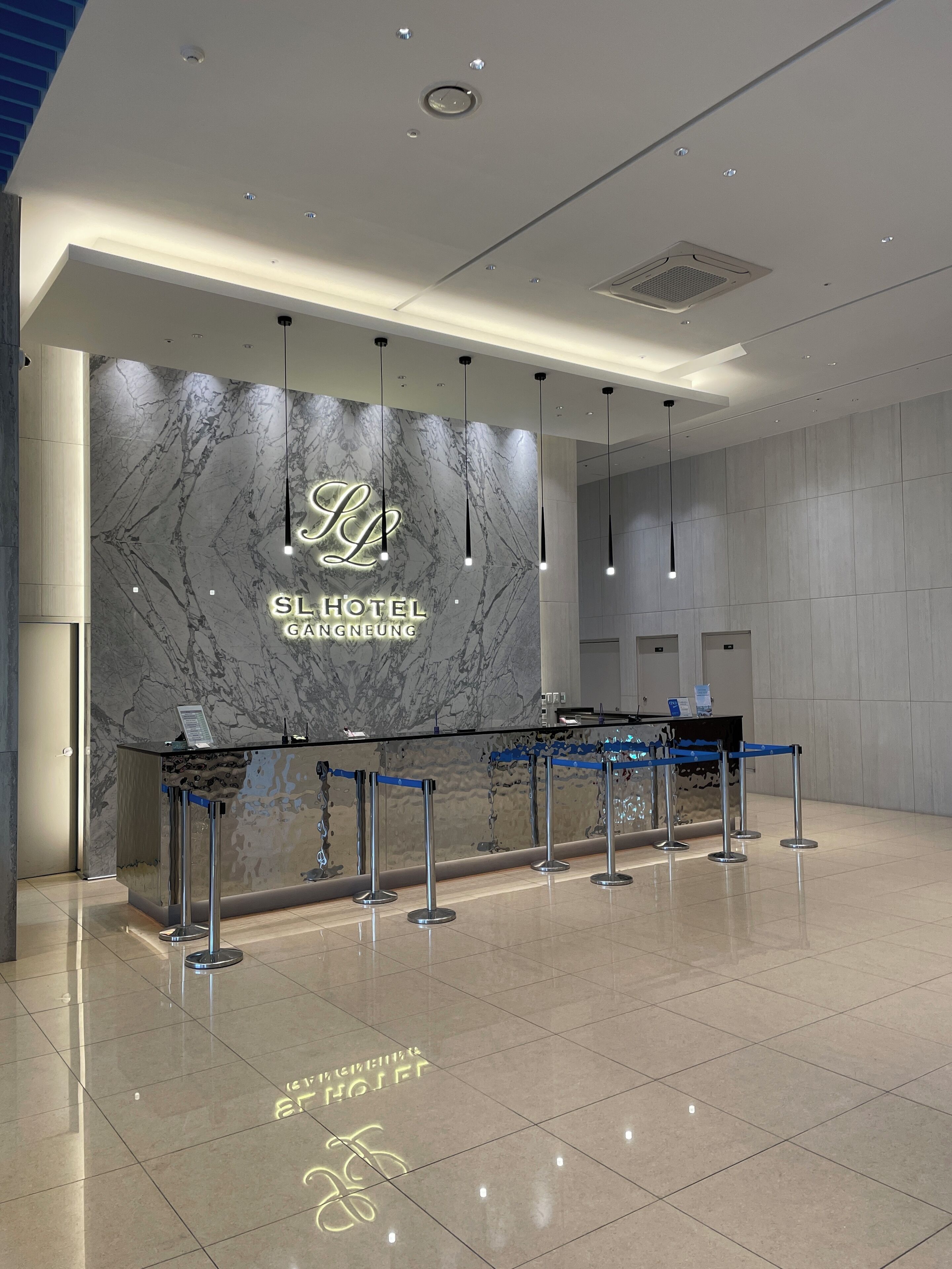 Zona de recepción de hotel con pared de mármol, mostrador de recepción y una barrera azul para organizar la fila.