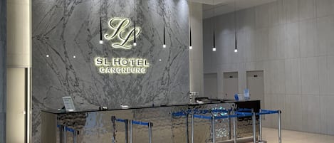 Zona de recepción de hotel con pared de mármol, mostrador de recepción y una barrera azul para organizar la fila.
