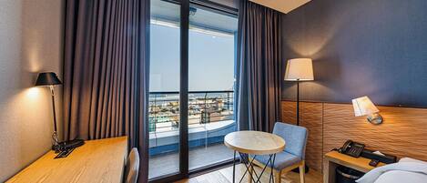 A hotel room with a wooden desk, a small round table, a chair, a bed, a nightstand, a lamp, and a view of the city through large windows.