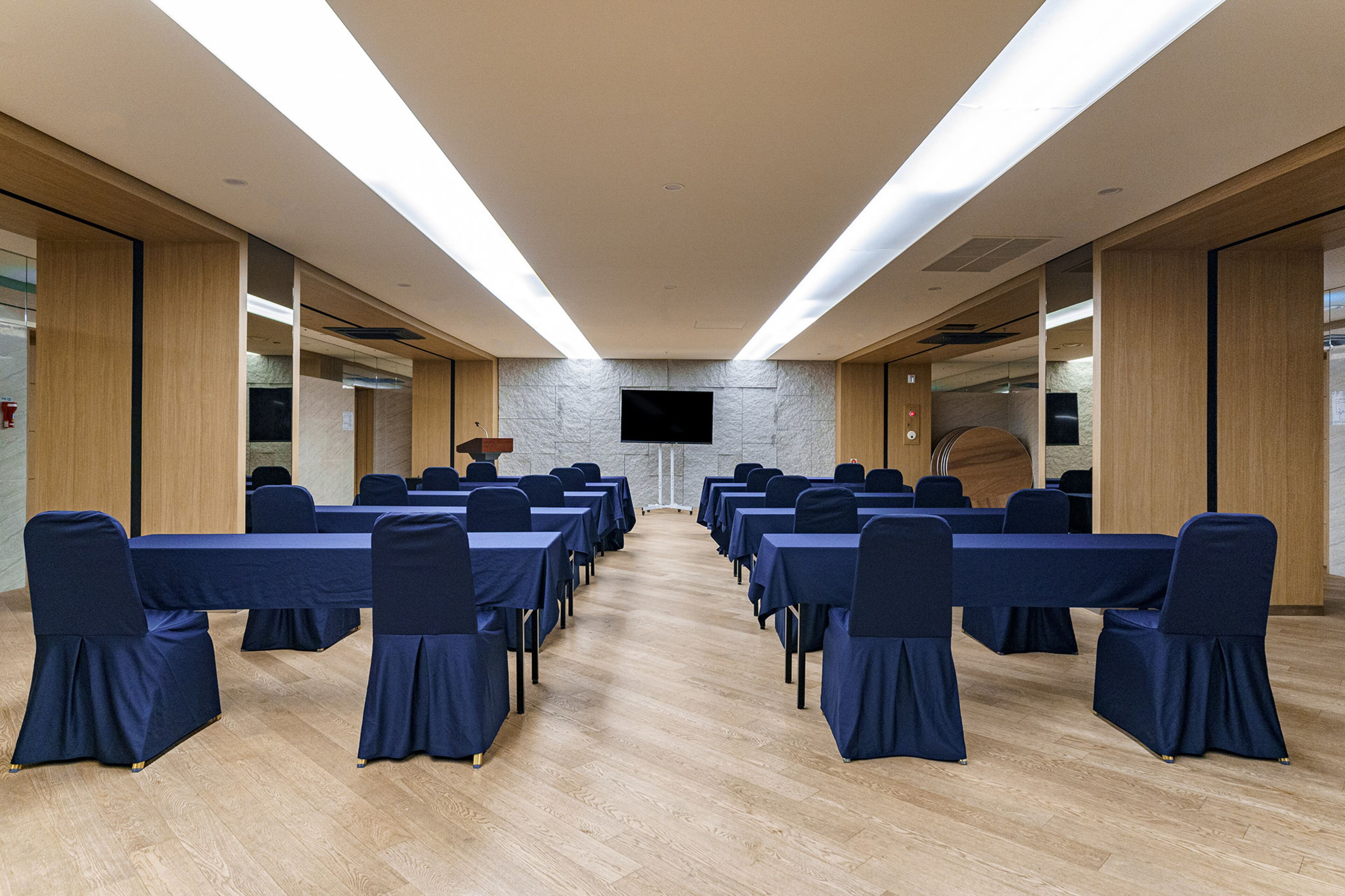 Ein Konferenzraum mit langen Tischen und Stühlen, einem Flachbildfernseher an der Wand und Holzvertäfelung.