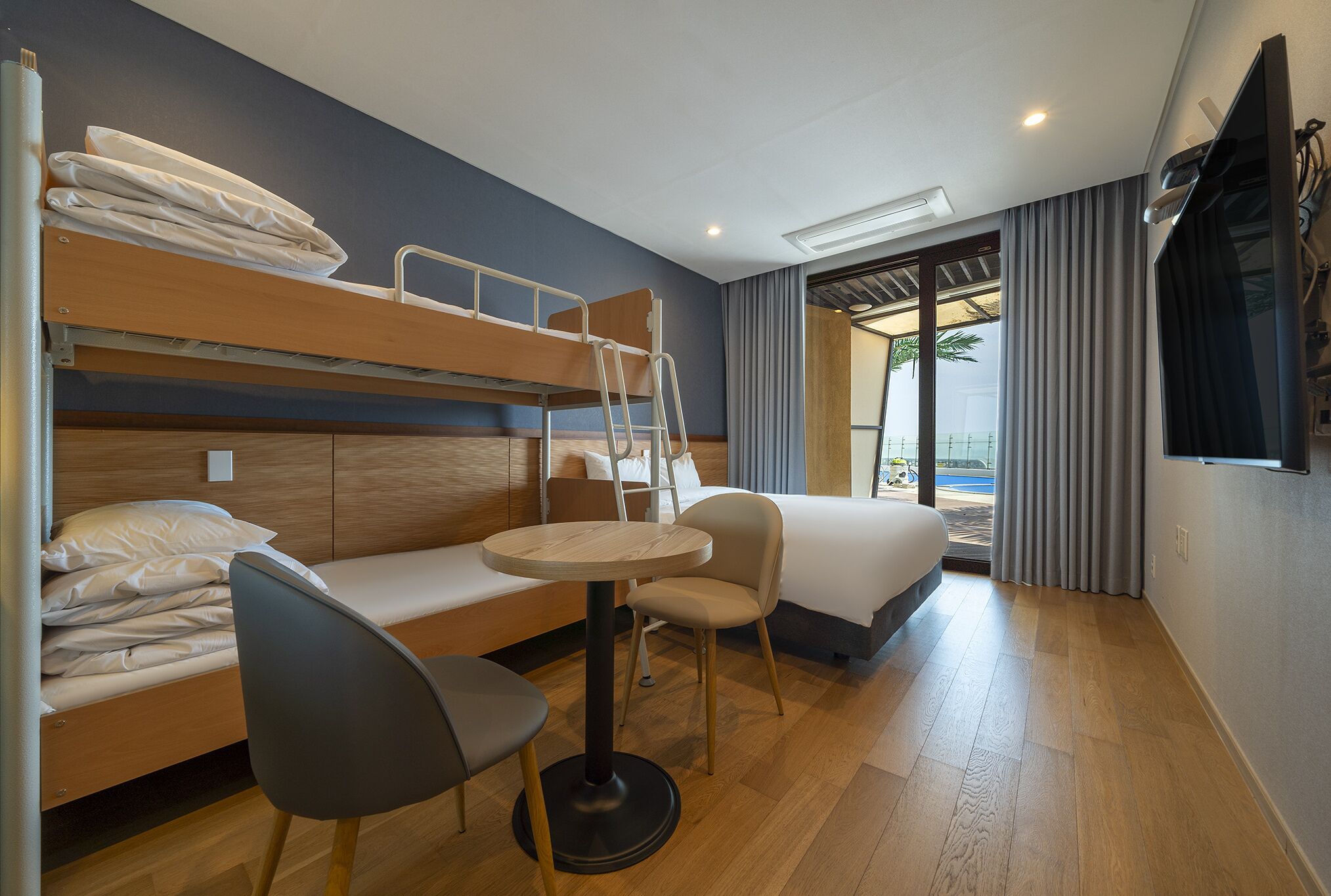 Habitación de hotel con literas, una mesita redonda pequeña, una silla, una cama, un televisor y una ventana con cortinas.
