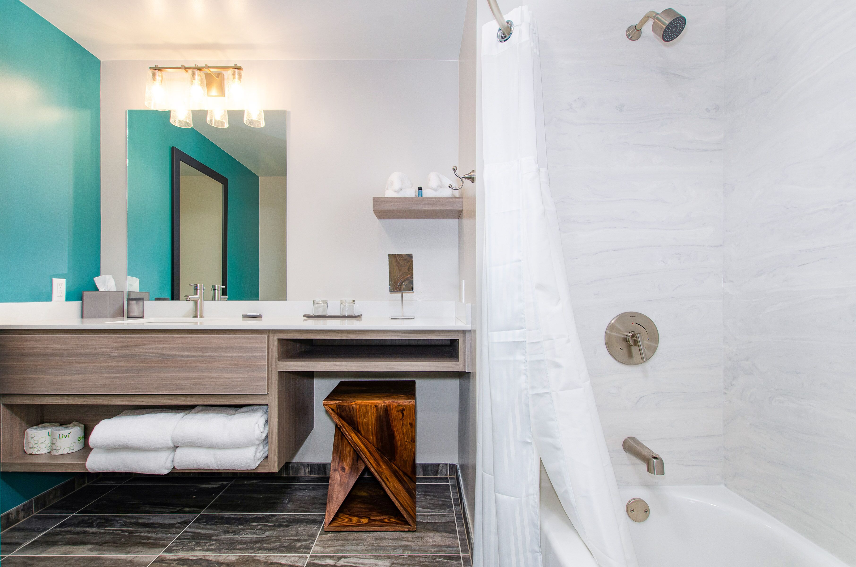A modern bathroom with a large mirror, a vanity with a wooden base, a shower with a curtain, and a small wooden stool.