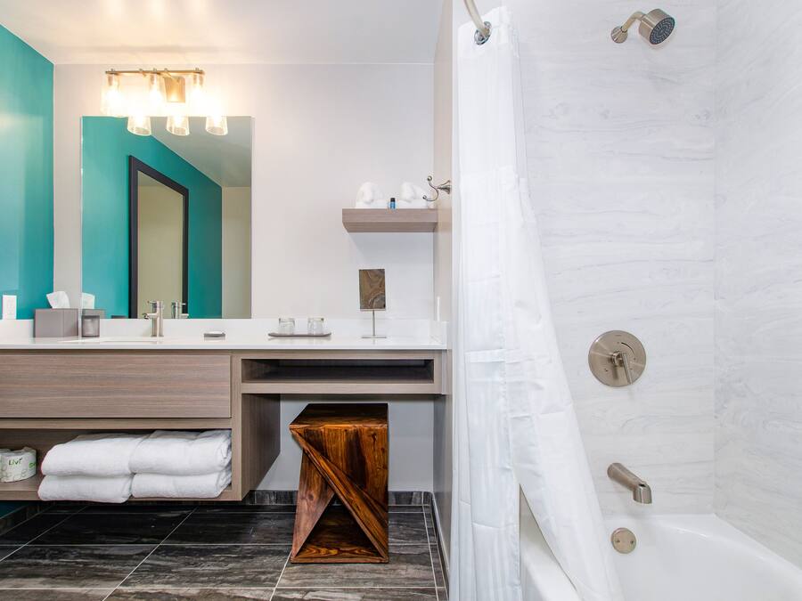 A modern bathroom with a large mirror, a vanity with a wooden base, a shower with a curtain, and a small wooden stool.