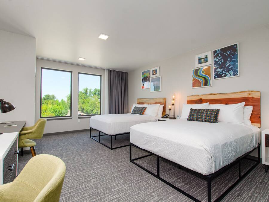 Habitación de hotel con dos camas, un escritorio, una silla y ventanas amplias con vistas a zonas verdes.