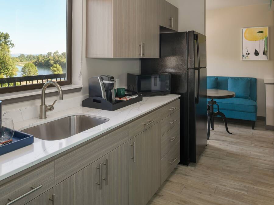 Una cocina moderna con un lavamanos, cafetera, microondas y refrigerador. Se ve una ventana con vista a árboles y un lago.
