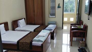 A hotel room with three beds, a wooden wardrobe, a desk with chairs, a television, and a window with a view of trees.