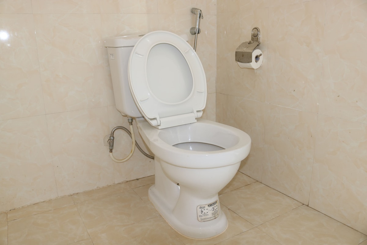Inodoro blanco estándar con palanca de descarga y un accesorio bidé en un baño con paredes de azulejos color beige.