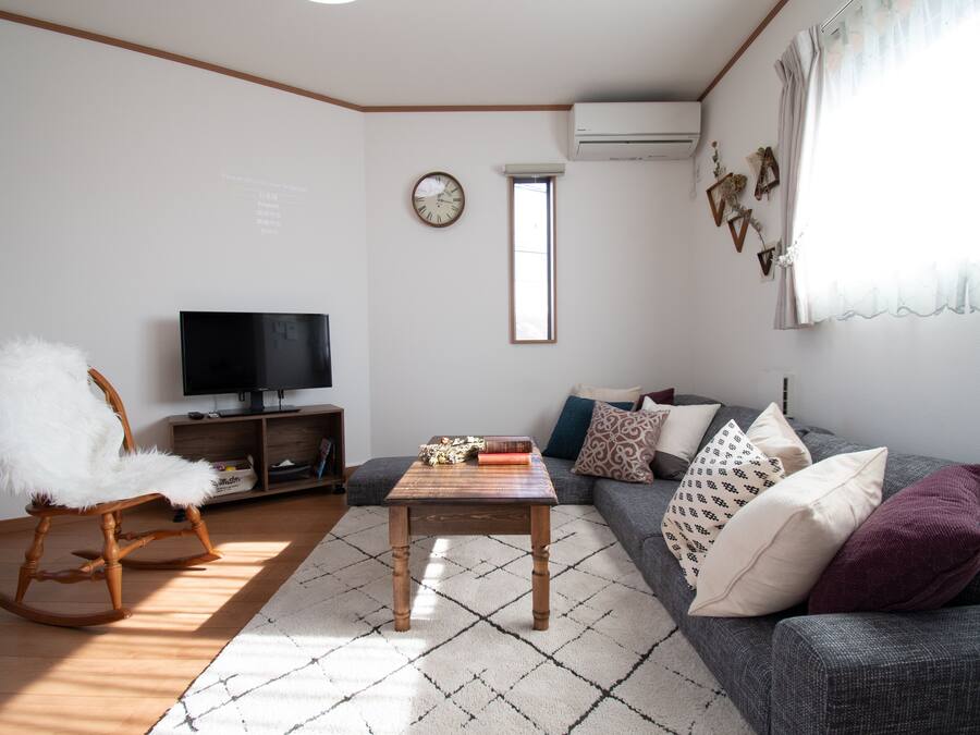 A living room with a wooden rocking chair, a television, a grey sofa with patterned cushions, and a small table with books.
