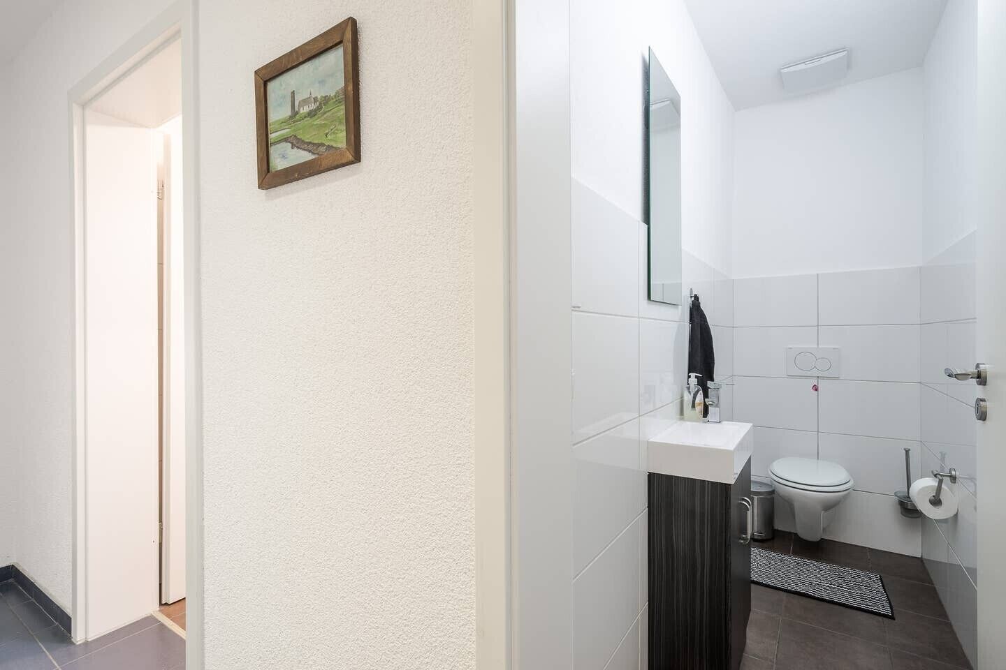 A bathroom with a white sink, toilet, and a mirror. There is a small rug on the floor and a picture frame on the wall.