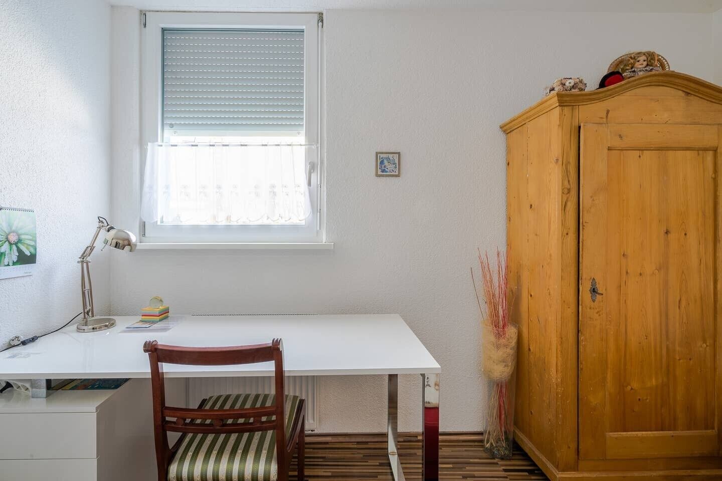 A room with a white desk, a wooden chair, a wooden wardrobe, and a window with blinds.
