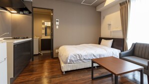 Habitación de hotel moderna con cama, sofá, pequeña mesa, kitchenette y baño visible a través de una puerta abierta.