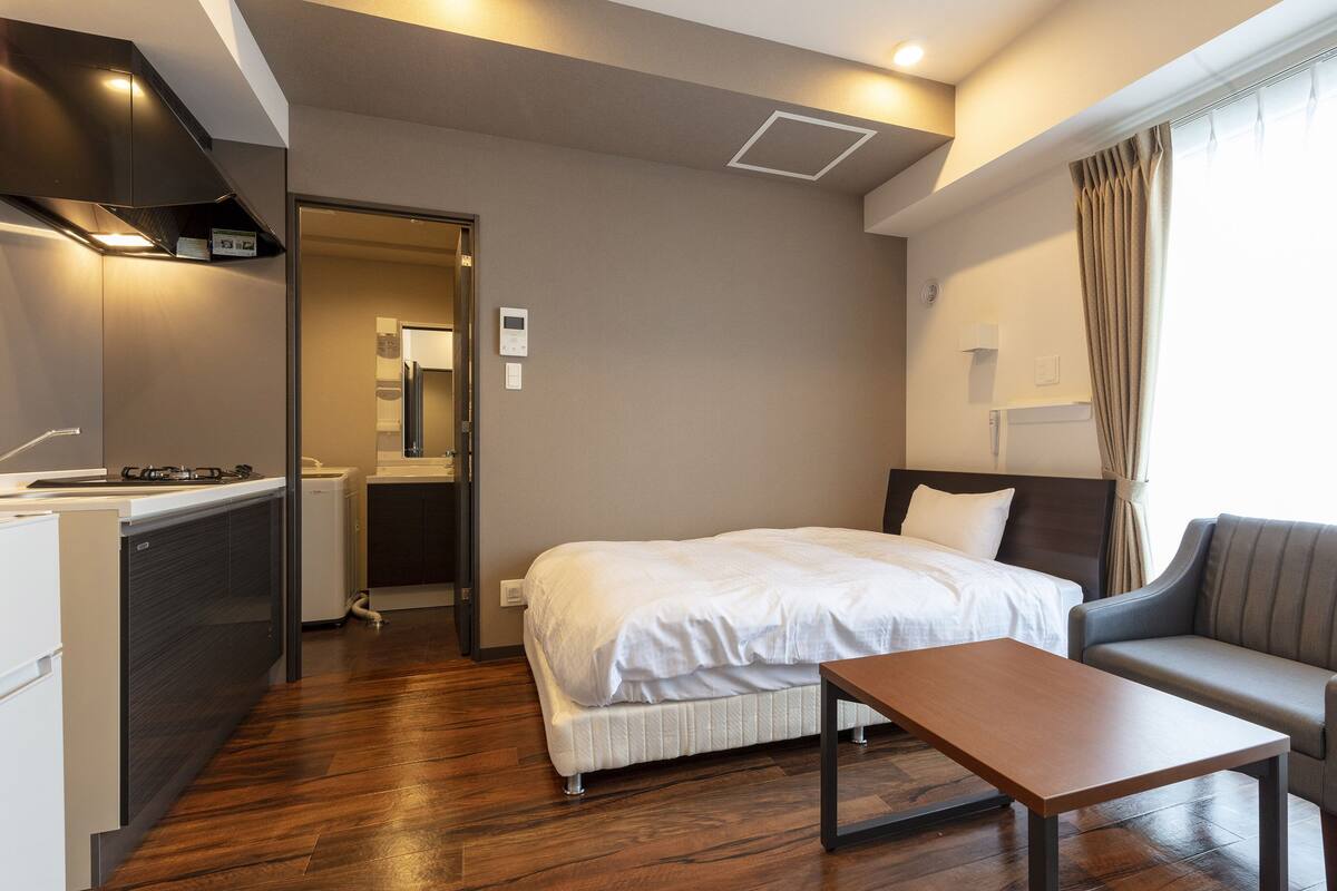A modern hotel room with a bed, a sofa, a small table, a kitchenette, and a bathroom visible through an open door.