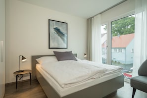 A modern bedroom with a bed, a small table with a lamp, a framed picture on the wall, and a view of the outside.