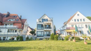 Ein mehrgeschossiges Gebäude mit Balkonen, ein Café namens 'STRAND-CAFE' und eine Rasenfläche mit Topfpflanzen.