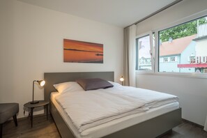 A neatly made bed with a pillow, a bedside lamp, a small table, and a window with a view of buildings. - Holiday Apartment "Montfort" Directly on Lake Constance with Views of the Lake and Mountains (Langenargen)