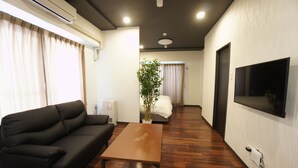 A living room with a black sofa, a wooden coffee table, a potted plant, a bed with white bedding, and a mounted TV. - Piece Eniwa (Eniwa)