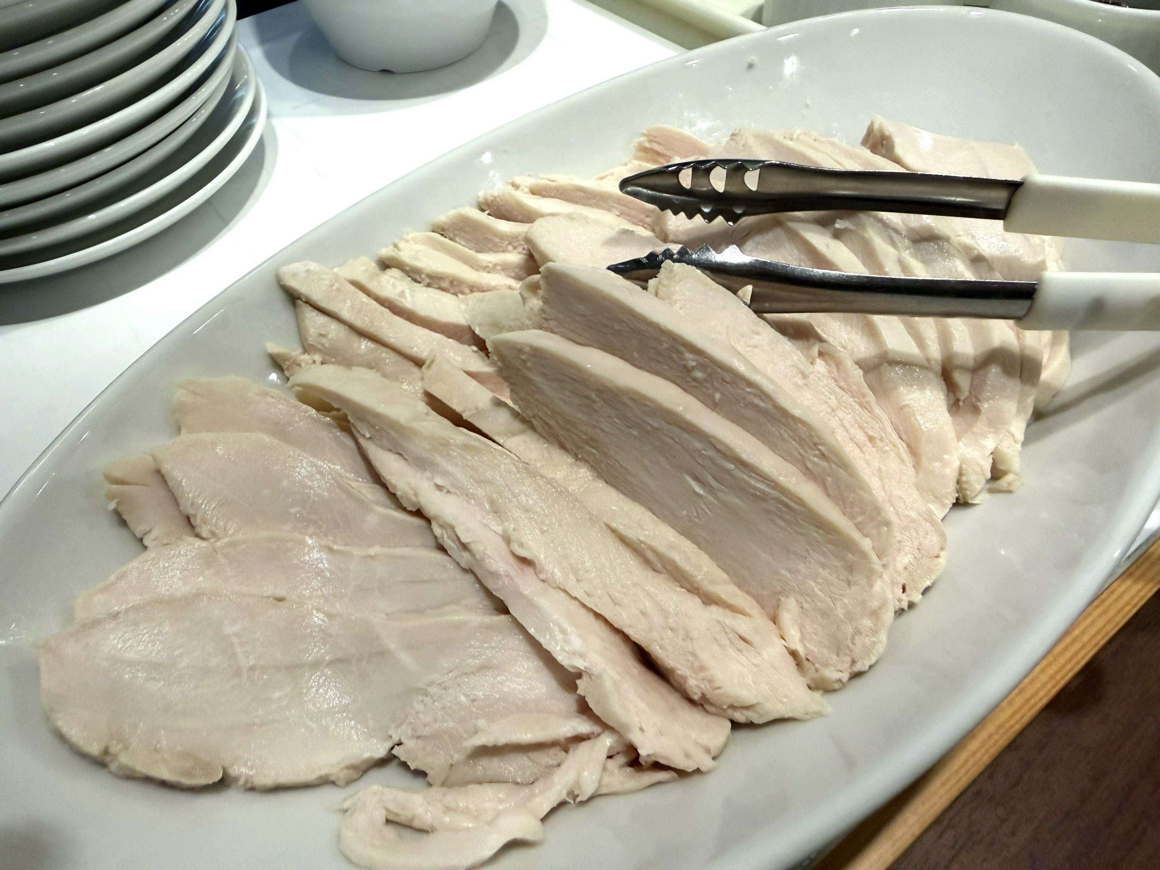 Un plato blanco con pechuga de pollo en rodajas, unas pinzas y una pila de platos al fondo.